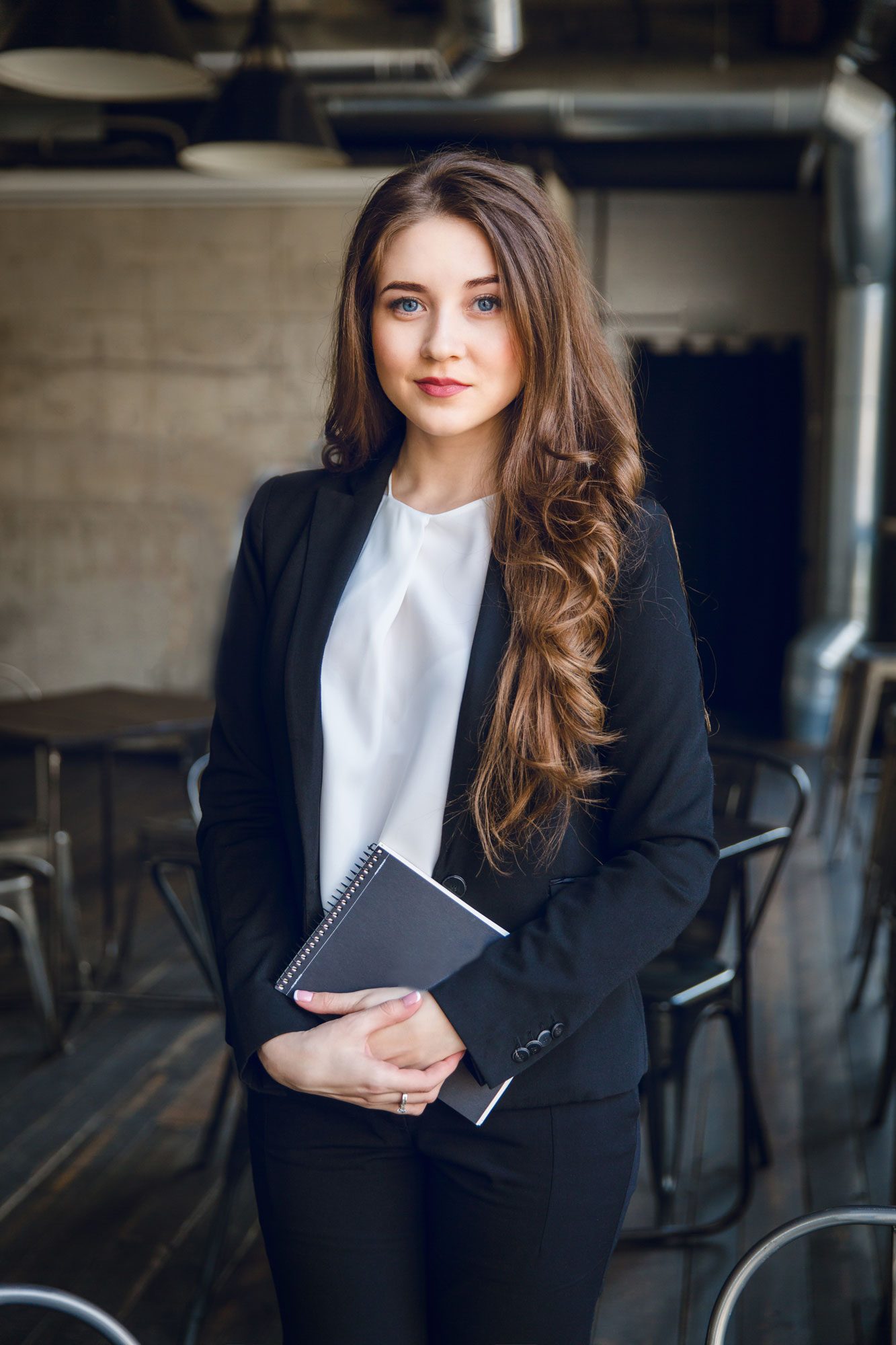 brunette-business-woman-with-wavy-long-hair-blue-eyes-stands-holding-notebook-hands-min
